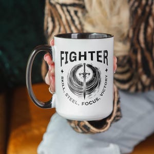 May include: White ceramic mug with a black interior and handle. The mug features the word "FIGHTER" above a sword design, with the words "SKILL, STEEL, FOCUS, VICTORY" surrounding the design. The mug is held by a person.