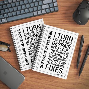 two spiral notebooks with humorous text printed on them, placed on a wooden surface alongside a computer keyboard, mouse, and other office supplies.