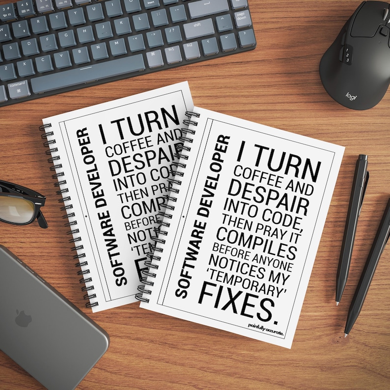 two spiral notebooks with humorous text printed on them, placed on a wooden surface alongside a computer keyboard, mouse, and other office supplies.