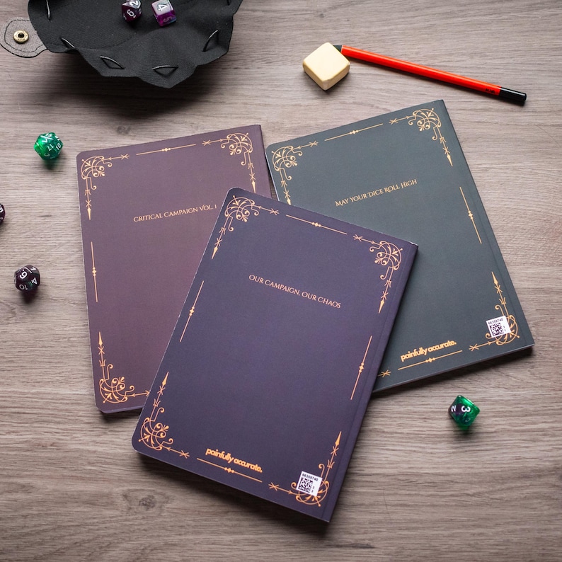 three books with gold-colored borders and text, along with a red pen and some dice on a wooden surface.