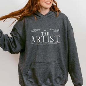 May include: A dark heather gray hooded sweatshirt featuring white text that reads A WORLD OF MAGIC THE PROBLEM SOLVER TECH ARTIST WHERE ART MEETS CODE—CREATING THE IMPOSSIBLE, DEBUGGING THE IMPROBABLE, AND MAKING IT LOOK EFFORTLESS.