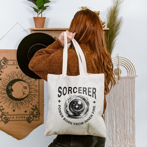 May include: Cream-colored tote bag with the word "SORCERER" in black, along with a circular graphic and the text "POWER BORN FROM INNER FIRE." The bag has two handles and is held by a person wearing a brown sweater.