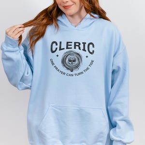 Puede incluir: Sudadera con capucha azul claro con bolsillo delantero y capucha con cordón. El gráfico negro en la parte delantera dice "Cleric" encima de un diseño de martillo y diana, con el texto "One prayer can turn the tide" alrededor.