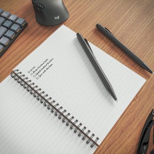 a wooden desk with a spiral-bound notebook, a black pen, a black keyboard, and a black mouse.