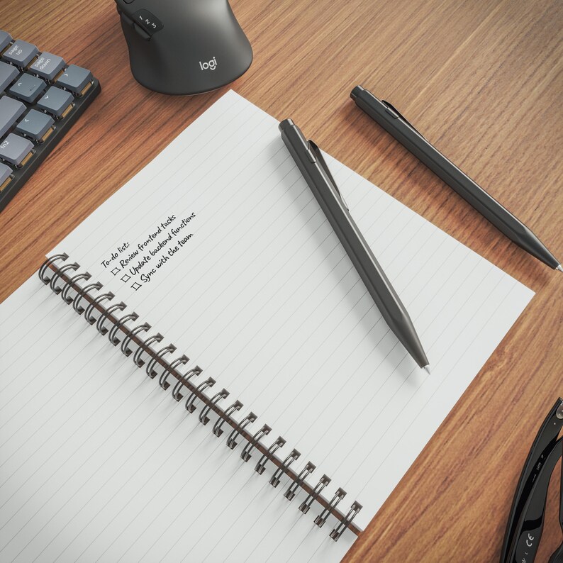 a wooden desk with a spiral-bound notebook, a black pen, a black keyboard, and a black mouse.
