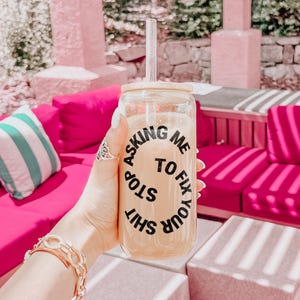 May include: A clear glass tumbler with a bamboo lid and a straw, filled with a light-coloured beverage. The tumbler has the text "Asking me to stop fix your shit" in black. The background features pink couches and a striped pillow.