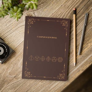 a brown campaign journal with gold decorative elements, placed on a wooden surface alongside a camera and a pencil.