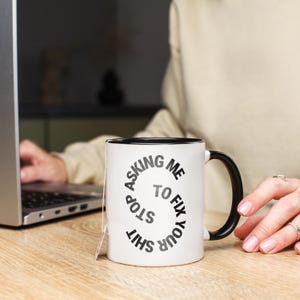 May include: White ceramic mug with a black interior and handle. The mug features the text "STOP ASKING ME TO FIX YOUR SHIT" in a spiral design. A tea bag is attached to the mug. The mug is on a wooden surface.