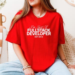 May include: A bright red t-shirt featuring the white text "I'm Just A FULLSTACK DEVELOPER EST 2013". The shirt has short sleeves and a round neckline. The text is centered on the front of the shirt.
