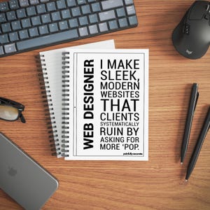 May include: A notebook with the text "WEB DESIGNER" and "I MAKE SLEEK, MODERN WEBSITES THAT CLIENTS SYSTEMATICALLY RUIN BY ASKING FOR MORE 'POP." is on a wooden desk. A keyboard, mouse, pens, phone, and glasses are also on the desk.