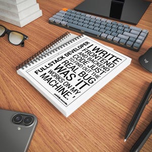a desk with various office supplies, including a spiral notebook, a keyboard, a mouse, and a pair of glasses. The notebook has a quote printed on it, which reads "I write frontend and backend code. I find out the real bug in my machine."