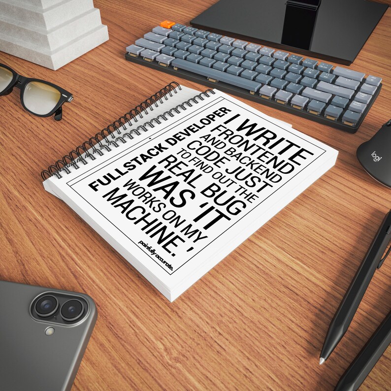 a desk with various office supplies, including a spiral notebook, a keyboard, a mouse, and a pair of glasses. The notebook has a quote printed on it, which reads "I write frontend and backend code. I find out the real bug in my machine."