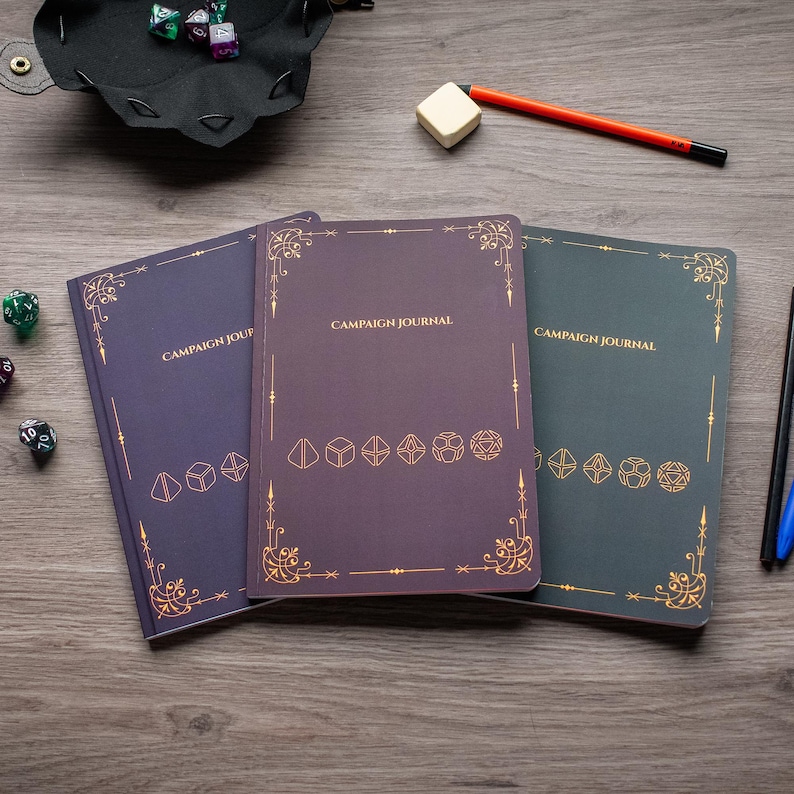 Three campaign journal notebooks with gold designs and text, along with a black pen, a black and blue pen, and some dice on a wooden surface.