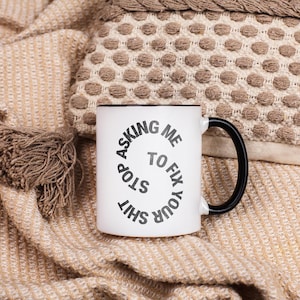 May include: White ceramic mug with a black handle and rim. The mug features the text "STOP ASKING ME TO FIX YOUR SHIT" in a circular design. The mug is set against a neutral-toned background.
