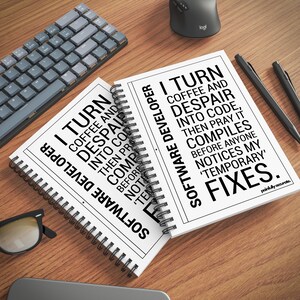 two spiral notebooks with humorous text printed on them, placed on a wooden desk alongside a keyboard, a mouse, and other office supplies.
