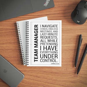 Könnte beinhalten: Eine weiße Karte mit schwarzem Text, der vertikal auf der linken Seite "TEAM MANAGER" lautet. Der Text der Karte lautet: "I NAVIGATE CHAOS, ENDLESS MEETINGS, AND LAST-MINUTE REQUESTS. ALL WHILE PRETENDING I HAVE EVERYTHING UNDER CONTROL." Ein Spiralnotizblock, Stifte und eine Computermaus sind ebenfalls sichtbar.
