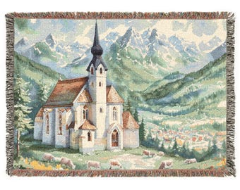 May include: A woven throw blanket featuring a detailed landscape scene of a white church with a tall steeple, situated in a green valley with grazing sheep in the foreground and snow-capped mountain peaks in the background, finished with fringe.