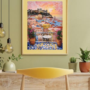 K&ouml;nnte beinhalten: Ein gerahmter Kunstdruck mit einem &Ouml;lgem&auml;lde eines mediterranen Balkons mit Blick auf eine K&uuml;stenstadt mit einer Burg auf einem H&uuml;gel bei Sonnenuntergang. Die Szene zeigt rosa Blumen in T&ouml;pfen, einen kleinen runden Tisch und einen blau-gelben Fliesenboden.
