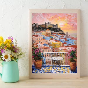 K&ouml;nnte beinhalten: Ein gerahmtes &Ouml;lgem&auml;lde, das einen mediterranen Balkon mit Blick auf eine Stadt mit bunten Geb&auml;uden und eine Burg auf einem H&uuml;gel bei Sonnenuntergang zeigt. Ein kleiner runder Tisch mit einem Weinglas steht auf dem gefliesten Balkon.