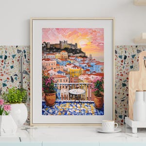 K&ouml;nnte beinhalten: Ein gerahmter Kunstdruck mit einem &Ouml;lgem&auml;lde einer mediterranen Stadt am Hang bei Sonnenuntergang. Die Szene zeigt eine Burg auf einem H&uuml;gel mit Blick auf bunte Geb&auml;ude, einen Balkon mit einem kleinen Tisch, zwei Topfpflanzen und einen gefliesten Boden.
