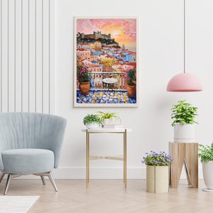 K&ouml;nnte beinhalten: Ein gerahmter Wandkunstdruck, der ein lebhaftes mediterranes Stadtbild bei Sonnenuntergang zeigt, mit einem Balkon, einem kleinen Tisch, Topfpflanzen und einem gefliesten Boden mit Blick auf bunte Geb&auml;ude und eine Burg auf einem H&uuml;gel.