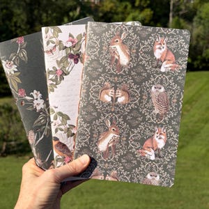 May include: Three rectangular notebooks with distinct covers. One features a dark background with white flowers and a bird. Another has a light background with floral patterns. The third displays a dark green repeating pattern of deer, foxes, and owls.