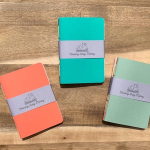 May include: Three colorful notebooks: coral, teal, and mint green, each wrapped with a gray band. The band features a logo with an open book, a heart, and the text "Chasing Daily Memory". The notebooks are on a wooden surface.