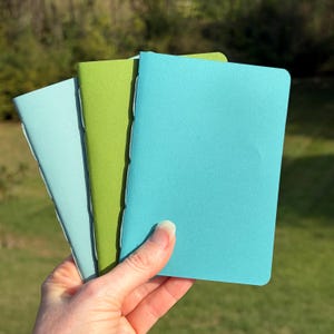 May include: Three small notebooks with solid-colored covers in shades of light blue, lime green, and bright turquoise. Each notebook features a simple rectangular shape with rounded corners and visible stitching along the spine edge.