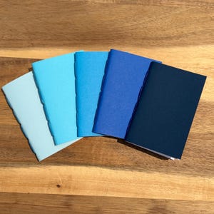 May include: A set of five rectangular notebooks arranged in a fan shape, featuring a gradient of blue covers ranging from a pale sky blue to a deep navy blue. Each notebook has a visible stitched spine along the left edge with a smooth finish.