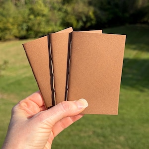 May include: Three small, rectangular notebooks with plain brown cardstock covers. Each notebook is bound with visible white stitching along the spine, featuring a simple, unadorned design with a matte texture and clean, sharp edges.