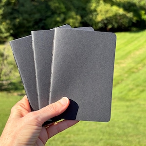 May include: Three small, rectangular notebooks with solid dark gray covers. Each notebook features a simple, clean design with rounded corners and a stitched spine, presented in a stacked arrangement to show the uniform matte texture.