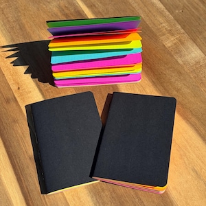 May include: A stack of neon-colored paper sheets with a dotted grid pattern, featuring shades of green, purple, orange, yellow, blue, and pink. Two black-covered notebooks are positioned in the foreground, resting below the colorful paper stack.