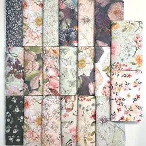 Op de afbeelding: Een verzameling van 18 kleine, vierkante notitieboekjes met bloemen- en vlinderontwerpen. De notitieboekjes zijn gerangschikt in drie rijen van zes. De omslagen zijn een mix van roze, groen, blauw en grijs.