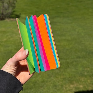 May include: A small, rectangular notebook with a spiral binding, featuring pages in vibrant neon colors including lime green, teal, magenta, orange, and cyan. Each page has a subtle dot grid pattern, and the pages are fanned out to show the spectrum.