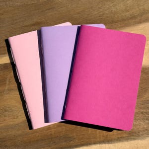 May include: Three rectangular notebooks with matte covers in shades of light pink, lavender, and magenta. Each notebook features a simple, clean design with visible black stitching along the spine, showcasing a minimalist aesthetic for note-taking.