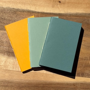 May include: Three small notebooks in shades of yellow, teal, and blue-green are arranged on a wooden surface. The notebooks have a simple, stitched binding. The covers are solid colors, and the pages are a light cream color.