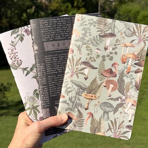 May include: Three rectangular notebooks with distinct covers. One features a light background with botanical illustrations, one is black with white printed text, and one displays a light green background with a detailed mushroom and fern pattern.