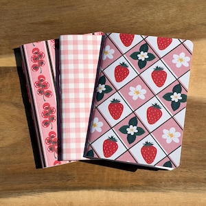 May include: Three notebooks with different covers on a wooden surface. One has a pattern of red tomatoes on a pink background, another has a pink gingham pattern, and the third features a strawberry design with white flowers and green leaves.