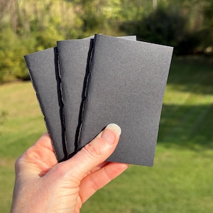 May include: Three small, rectangular, matte black paper booklets with visible stitched spines. Each booklet features a plain, solid black cover and a simple folded construction, presented as a set of three identical items.