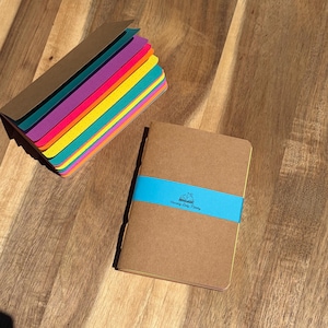 May include: A stack of small notebooks with brown covers and multicolored pages, alongside a single brown notebook wrapped in a bright blue paper band featuring the text Chasing Daily Money and a small book icon illustration.