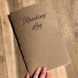 May include: A brown reading log notebook with the words "Reading Log" in cursive script. The notebook is lying on a wooden surface. A hand with a gold ring is holding the notebook.