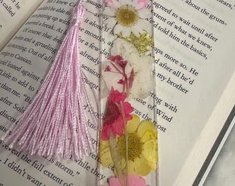 May include: A pink and yellow pressed flower bookmark with a pink tassel. The bookmark is made of clear resin and features a variety of pressed flowers, including daisies, roses, and wildflowers.