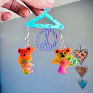 May include: A pair of tie-dye bear earrings hanging from a blue plastic hanger. The earrings are made of clear resin with a tie-dye pattern in shades of orange, yellow, green, blue, and pink. The bears are standing on their hind legs with their arms outstretched.