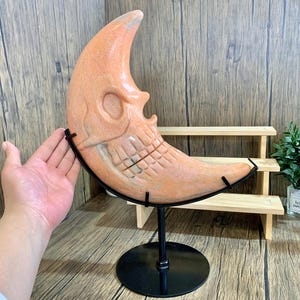 May include: A carved pink and orange crescent moon sculpture with a skull design. The sculpture is on a black metal stand with a round base.