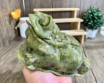 Könnte beinhalten: Eine grüne Jade-Froschskulptur mit einem detaillierten Design. Der Frosch sitzt auf seinen Hinterbeinen mit erhobenen Vorderbeinen. Der Frosch hat einen großen, runden Bauch und einen kleinen Kopf. Die Skulptur ist aus glattem, poliertem Jade gefertigt.