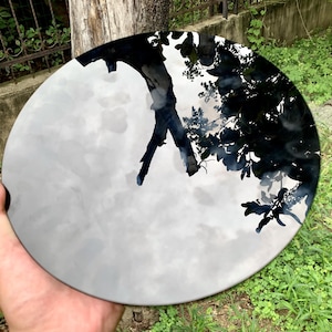 Peut inclure: Une œuvre d'art circulaire et réfléchissante représentant un arbre et un ciel. Le design présente une silhouette d'arbre noire sur un fond nuageux et gris. La pièce est tenue dans une main, avec de l'herbe verte en arrière-plan.