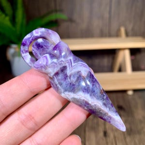 Può includere: Un cristallo di ametista viola e bianco scolpito in una forma appuntita con un anello nella parte superiore. Il cristallo ha un motivo a spirale ed è lucidato per una finitura liscia.