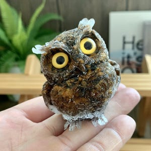 May include: A detailed figurine of an owl with large yellow eyes and a brown and white speckled body. The owl has a slightly tilted head and is perched on a surface. The figurine is a decorative item.