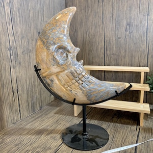 May include: A crescent moon-shaped sculpture carved from a light brown and gray stone with a skull-like face. The sculpture is mounted on a black metal stand with a round base.