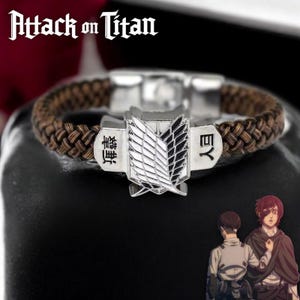 Puede incluir: Un brazalete de cuero marrón con un dije de metal plateado que presenta el logotipo de Ataque a los Titanes y texto japonés. El dije es un símbolo alado con el texto "翼" y "団" a cada lado.